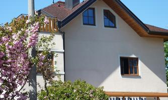 A charming house with a hipped roof and large windows. In front of the house, there are blooming trees and greenery.