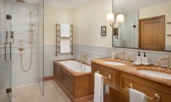 An elegant bathroom with a glass shower and a bathtub. The double sink is made of wood with golden fixtures.