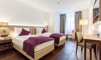 A modern room with two single beds, white bedcovers, and dark wooden floors. The room is brightly furnished and has a window with curtains as well as a small desk with a chair.