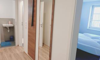 A bright hallway with a mirror and access to a bedroom. Nearby is a small bathroom with a sink.