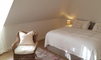 A bright bedroom with a cozy double bed and a rattan chair. Large windows provide plenty of natural light.