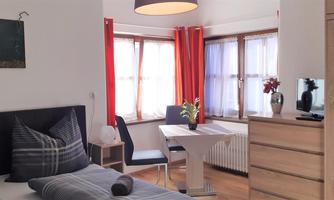 A cozy room with a double bed, a table, and chairs. Large windows provide plenty of daylight and a friendly atmosphere.