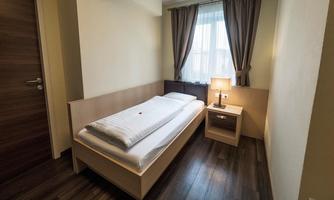 A simple bedroom with a single bed, a bedside table, and a window. The room has hardwood flooring and light walls.