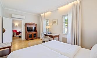 A bright bedroom with a double bed and a desk. The room has large windows and a cozy seating area.