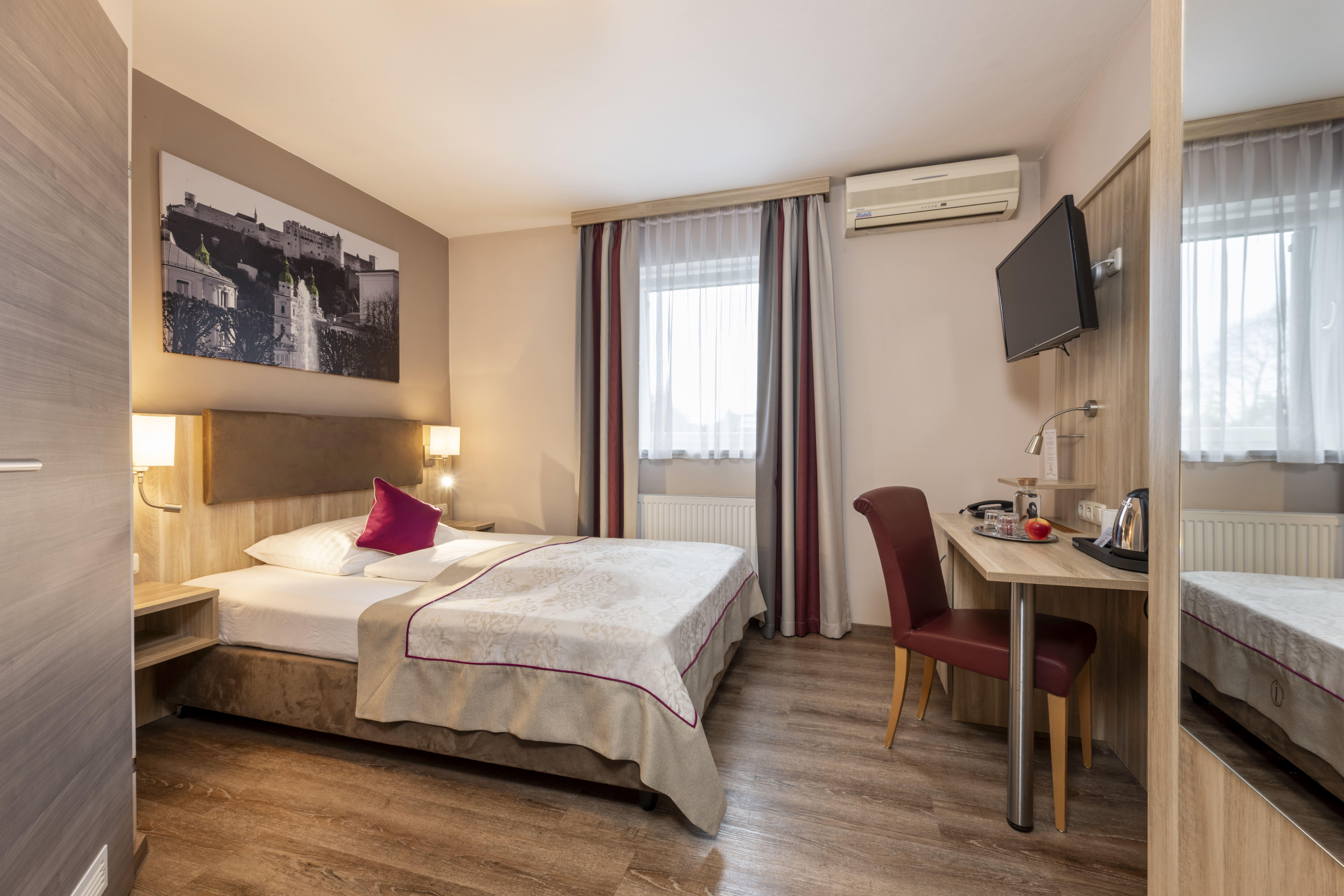 A cozy hotel room with a double bed, a desk, and a television. The windows are bright, and the walls are painted in warm tones.