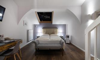 A modern hotel room with a double bed, a desk, and a television. The room has hardwood floors and large windows with daylight.