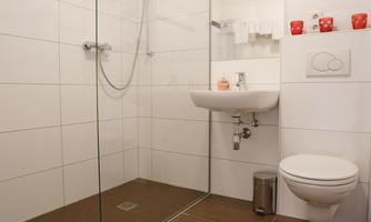 A modern bathroom with a glass shower cabin and light tiles. It includes a sink, a toilet, and practical shelves.