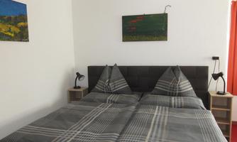 A modern bedroom with a double bed and a checkered duvet. Artworks hang on the walls, and the windows are decorated with orange curtains.