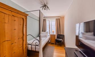 A cozy bedroom with a wooden wardrobe, a bed, and a television. Daylight streams through the windows, which are adorned with curtains.