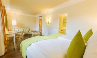 A bright and modern hotel room with a double bed and green pillows. In the background, a table and chairs as well as a colorful sofa can be seen.