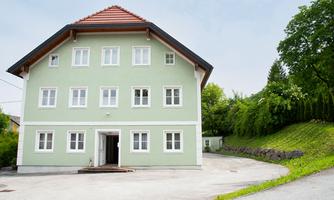 A green building with several windows and a red roof. It is located in a quiet environment with a lot of greenery.