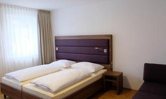 A simple, modern bedroom with a double bed and a cozy sofa. It has large windows that let in plenty of light.