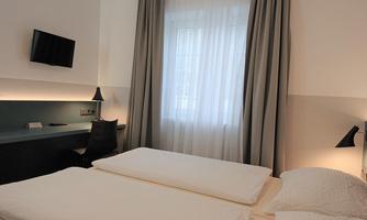A modern bedroom with two single beds and a large window. On the wall, there is a TV and a desk.