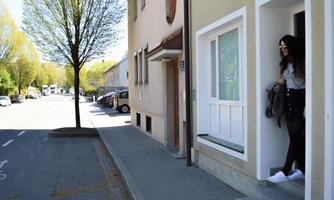 A woman stands at the door of a house and looks outside. The street is quiet and lined with trees.