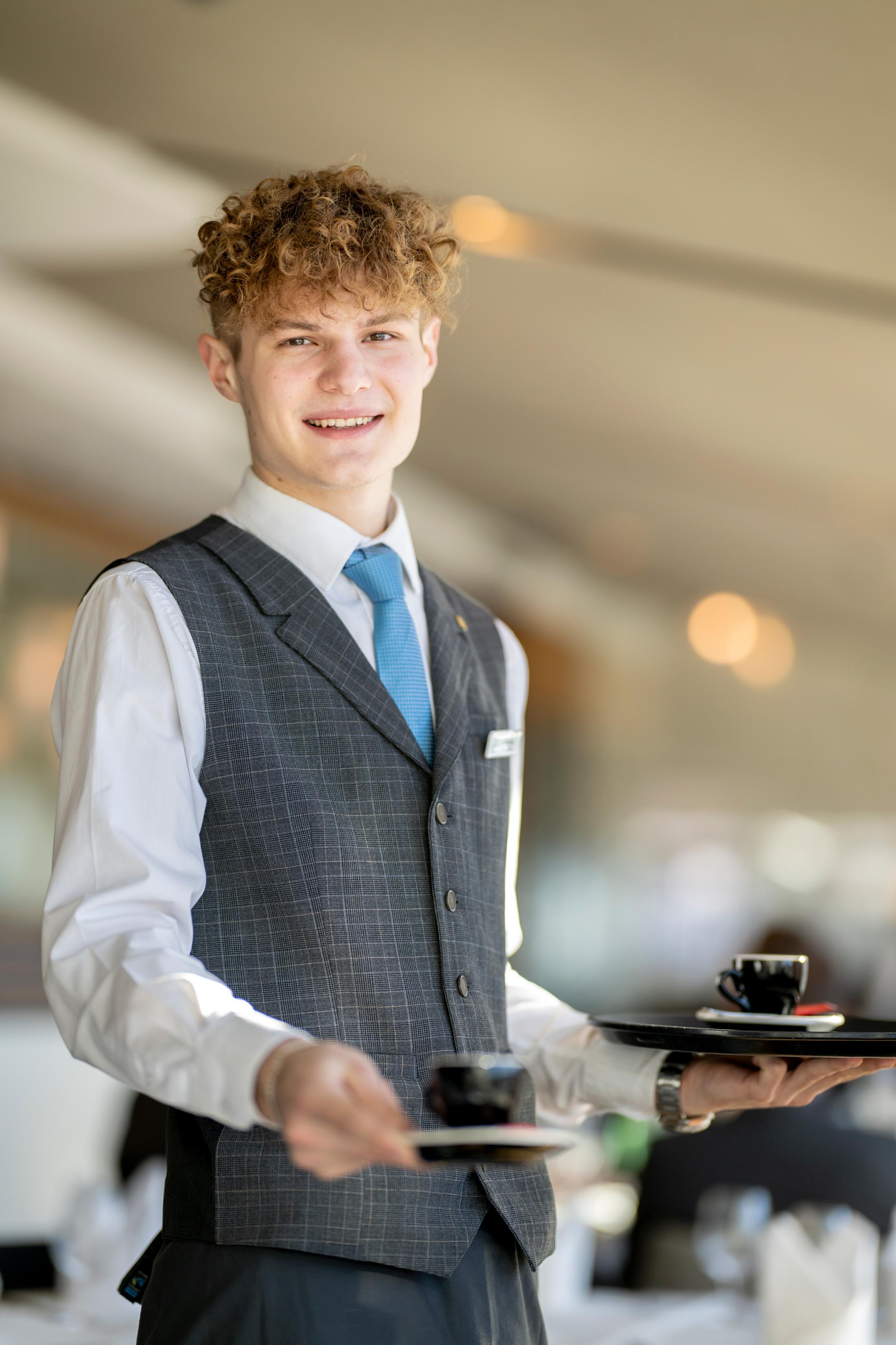 A young waiter is carrying two plates with cups. He is smiling kindly in an elegant restaurant.