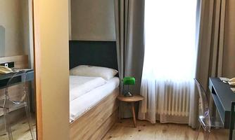 A simple room with a single bed, a table, and a chair. The window lets in plenty of light, and there is a large mirror.