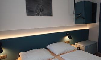 A modern bedroom with two beds and decorative wall lighting. A minimalist artwork hangs on the wall and there is a small side table.