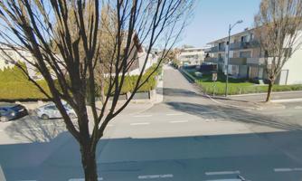 A quiet street with green areas and residential buildings on both sides. Some cars and trees are visible in the foreground.
