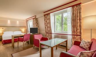 A modern hotel room with two beds, a table, and comfortable armchairs. Large windows let in plenty of natural light and offer a view of the greenery.