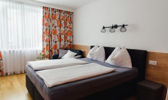 A modern bedroom with a large bed and elegant bedding. The windows are decorated with bright curtains, and the floor is made of wood.