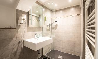 A modern bathroom with a walk-in shower and a simple sink. The walls are tiled in neutral gray tones, and there is heating as well as large mirrors.