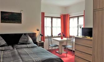 A cozy room with a double bed and large windows. The room is modernly furnished and has a small dining area with a table and chairs.