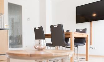 A modern dining area with a wooden communal table and elegant chairs. A large television is mounted on the wall.