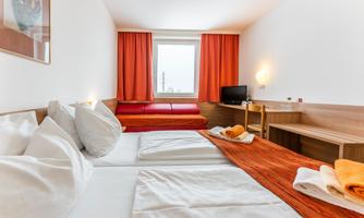 A simple hotel room with two beds, white bedspreads, and orange towels. Bright windows with orange curtains and a small desk.