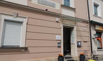 A bright building with an entrance and a distinctive canopy. In front of the door stands a flower pot, and on the left side, closed shutters can be seen.