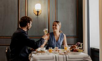 A couple is sitting at a table and clinking glasses. On the table, there are various breakfast dishes and drinks.