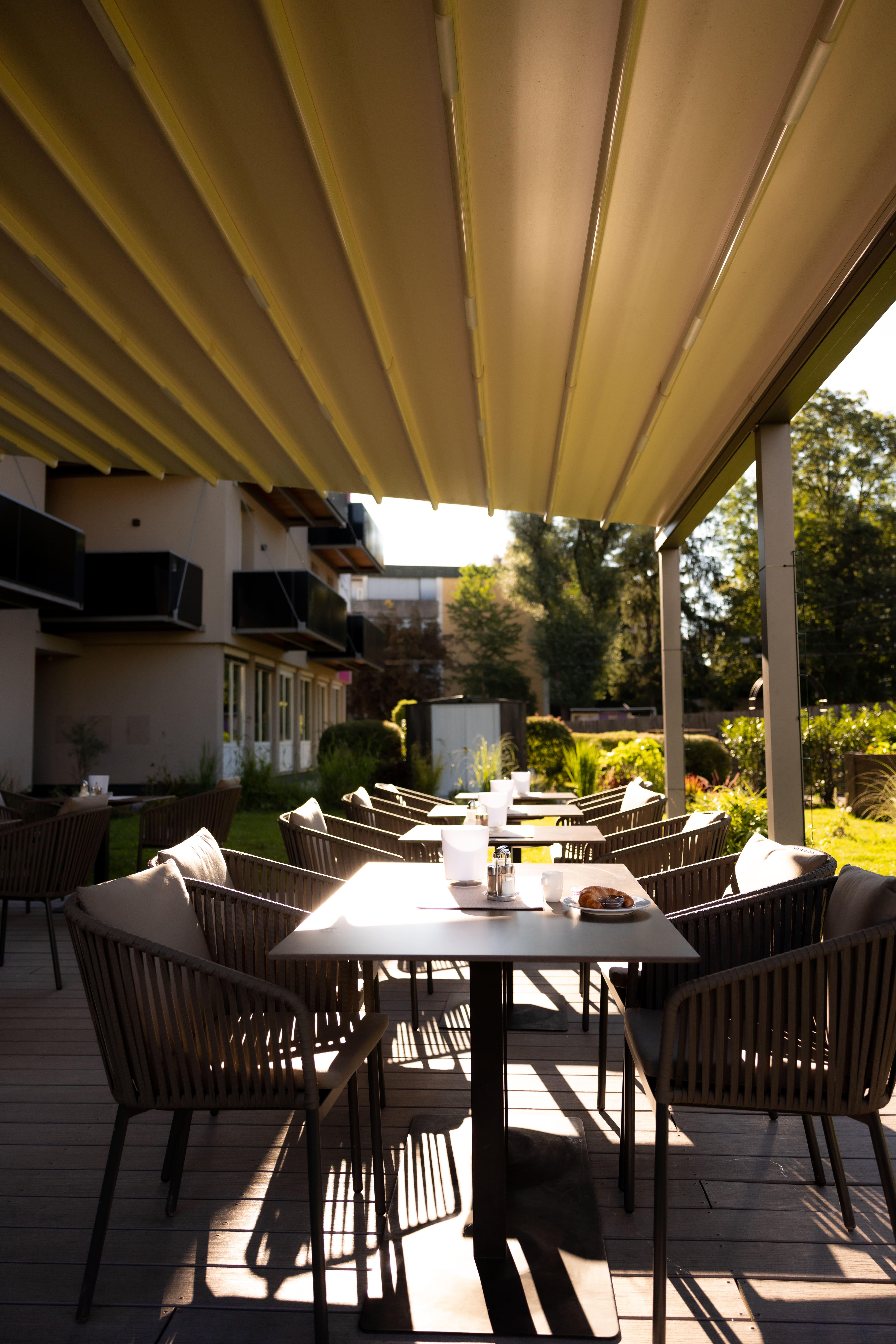 A cozy outdoor area with tables and chairs under sun protection. In the background, green plants and a building are visible.