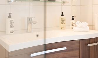 A modern bathroom with a white sink surface and wooden cabinets. On the sink, there are bottles and towels.
