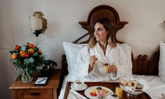 A woman is sitting in bed and enjoying a breakfast with various foods. Next to her is a bouquet of flowers, and she is wearing a bathrobe.