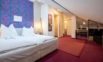 A cozy bedroom with a double bed and a purple accent wall. The room is bright and modernly furnished, with a small dining area.