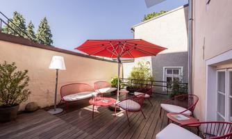 An inviting outdoor area with modern red furniture and a large sun umbrella. The wooden terrace is surrounded by plants and offers a cozy spot for relaxation.