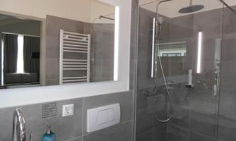 A modern bathroom with a glass shower and gray tiles. The mirror has integrated lighting, and there is a towel warmer in the background.