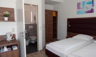 A modern room with two single beds and a wall unit. In the background, a bathroom and a small sitting area can be seen.