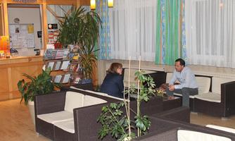 A cozy lounge with plants and comfortable seating. Two people are conversing in a warmly designed room.
