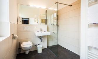 A modern bathroom with a glass shower, a sink, and a toilet. The walls are light-colored tiled and the floor is dark.