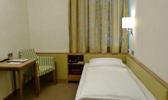 A simple room with a single bed, a table, and a chair. The room has a bright decoration with curtains and a wall lamp.
