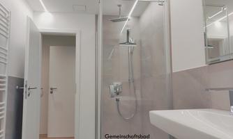A modern communal shower with a glass shower enclosure. In the background, bright walls and a sink are visible.