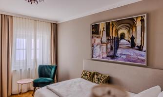 A cozy bedroom with a modern bed and a green armchair. A large picture of a historic interior hangs on the wall.