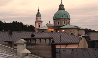 A city view with historic buildings and green domes. The sky is cloudy and the roofs are in various shades of gray.