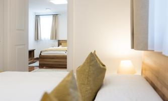 A cozy bedroom with a double bed and yellow pillows. Through the door, another room with a bed and windows is visible.