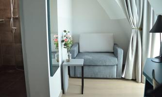 A cozy seating area with a gray sofa and a small table. A vase with flowers stands on the wall, and there is a curtain and a shower cabin in the background.