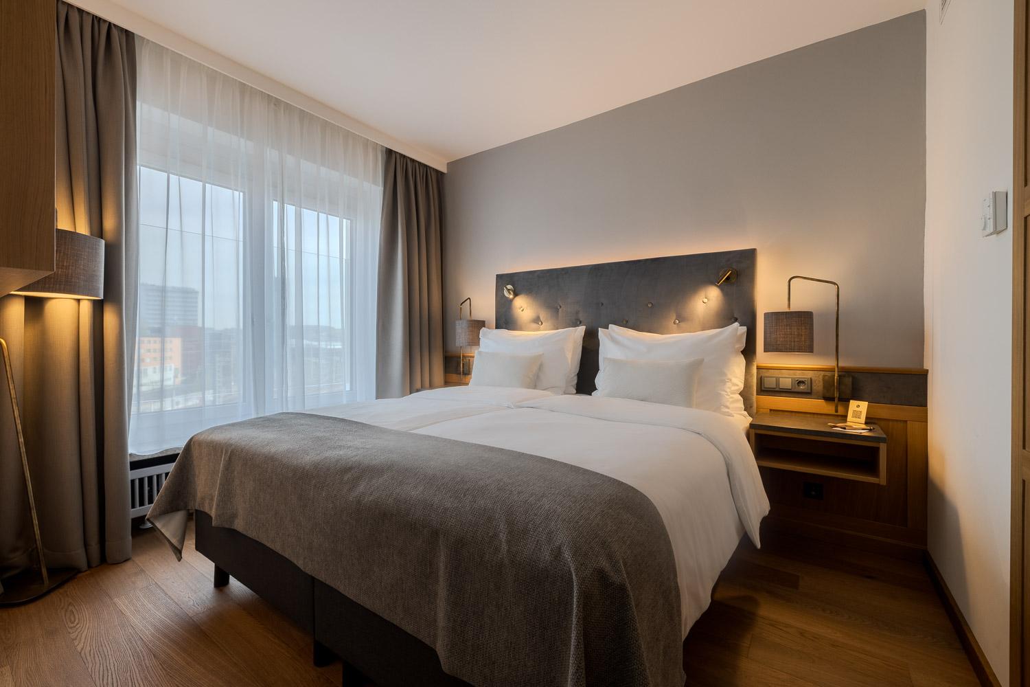 A modern hotel room with a double bed and soft color tones. Large windows provide plenty of daylight.