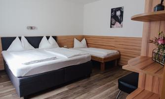 A simple bedroom with two beds, a modern kitchenette, and wooden paneling. The walls are light-colored and there is a decorative picture.