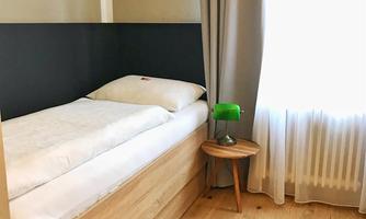 A simple bedroom with a single bed and a small nightstand. The room has light walls and a window with curtains.