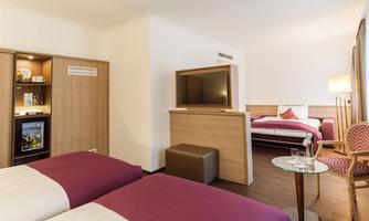 A modern hotel room with two single beds and an additional bed. There is a television, a minibar, and a table with chairs.