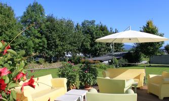 An inviting outdoor seating area with yellow furniture and a sunshade. The garden is green and sunny, surrounded by trees.